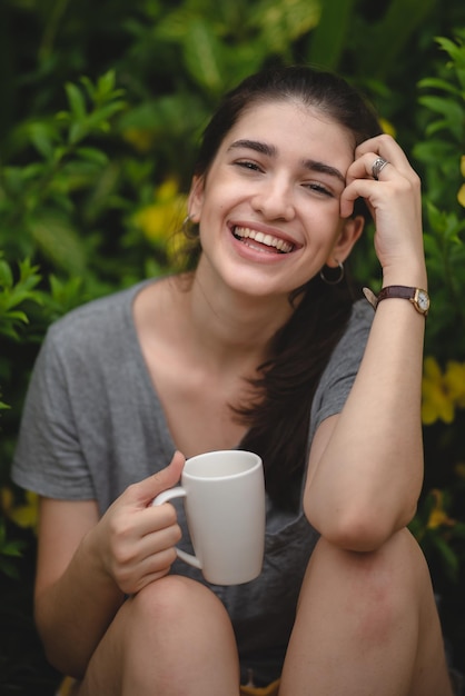 Cheerful young woman relaxing and breathing fresh air while sitting in balcony with nature view and drinking morning coffee and beverage