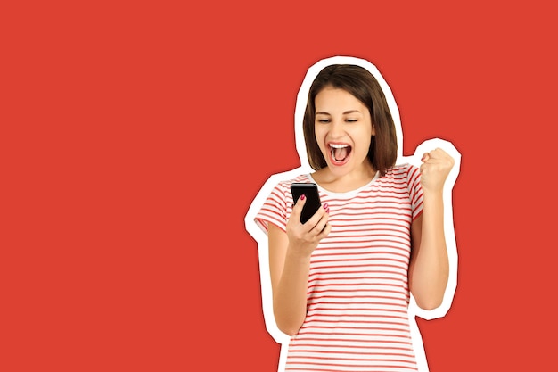 Cheerful young woman screaming while looking in mobile phone against red background