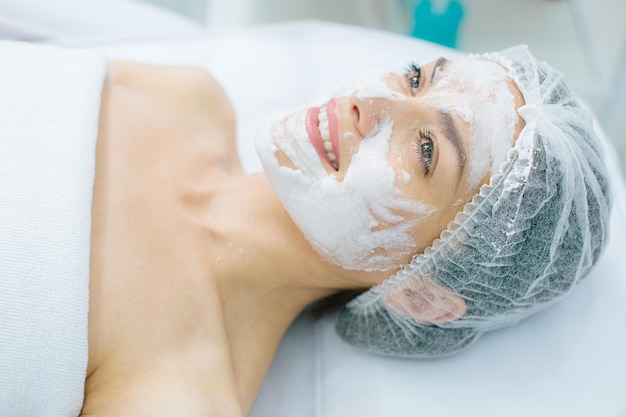 Cheerful young woman in a shower cap lying with oxygen mask on her skin and looking happy