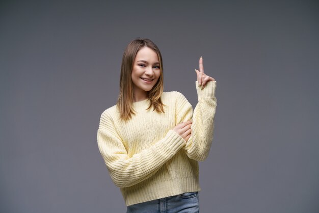 Cheerful young woman shows thumbs up in a yellow sweater on a gray wall, positive advertising