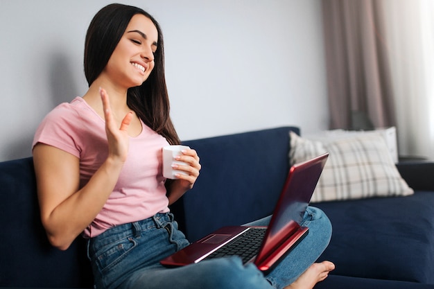 Cheerful young woman sit on sofa and wave with hand. She smiles and look on laptop screen. Model hold it on nap. She has white cup of drink.