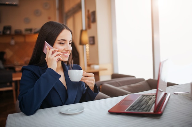 Cheerful young woman sit at table and work. She smiles. Customer talk on phone and look at laptop's screen. Young woman drink coffee from cup