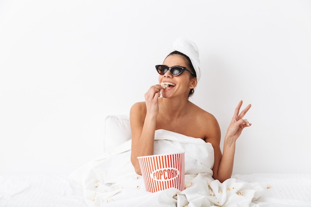 Allegra giovane donna seduta a letto dopo la doccia avvolta in una coperta, indossando occhiali da sole, mangiando popcorn