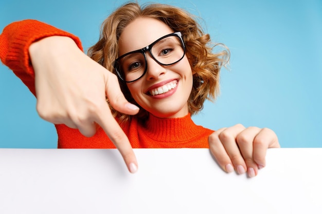 Cheerful young woman smiling for camera and pointing at empty banner against blue background