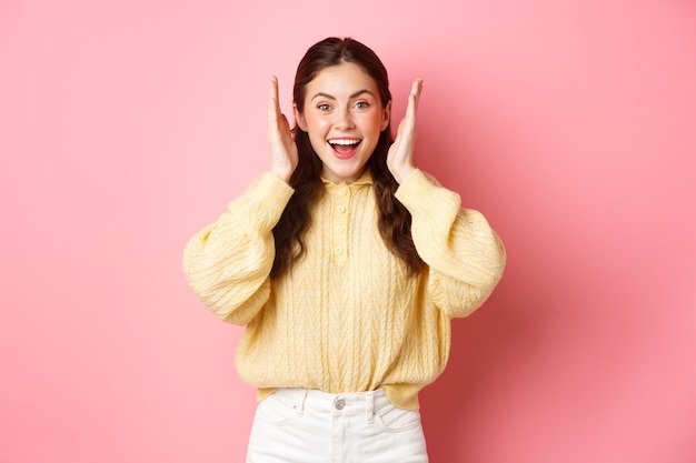 Cheerful young woman smiling, looking excited, open her ears after loud bang or fireworks, celebrating something, standing against pink bright wall