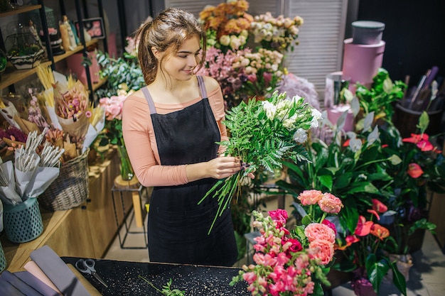 陽気な若い女性が立ち、花を組み合わせる。彼女は花束を作る。花屋の笑顔。彼女は花と植物でいっぱいの部屋に立っています。