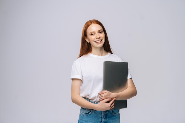 Cheerful young woman student holding laptop computer and looking at camera on isolated gray background. Pretty lady model with red hair emotionally showing facial expressions in studio, copy space.