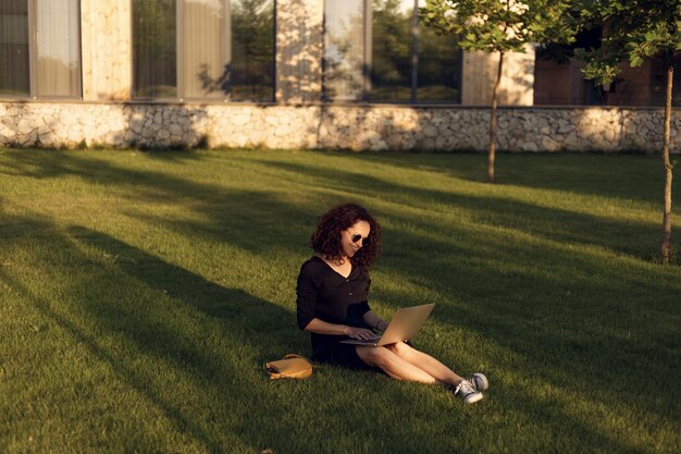 Cheerful young woman in sunglasses sitting on green grass during summer day while using laptop sitting on grass