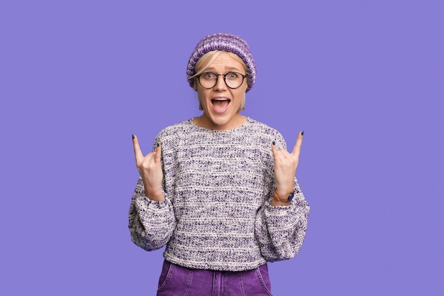 Cheerful young woman in sweater and hat isolated on purple wall background in studio Healthy fashion lifestyle cold season concept Depicting heavy metal rock sign