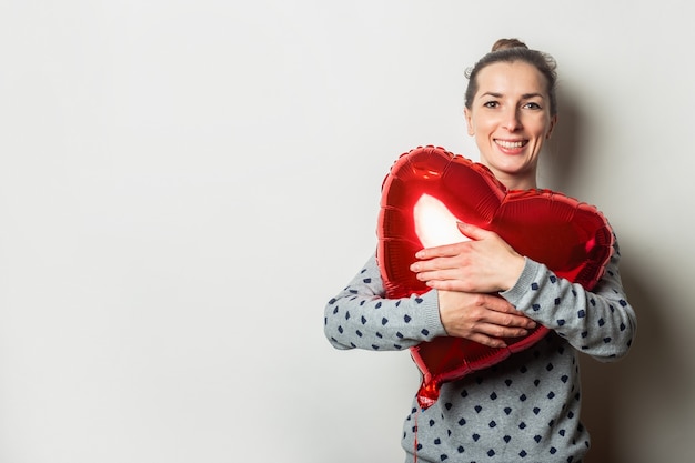 La giovane donna allegra in un maglione abbraccia un cuore dell'aerostato di aria su uno sfondo chiaro. concetto di san valentino. cerca una persona cara.