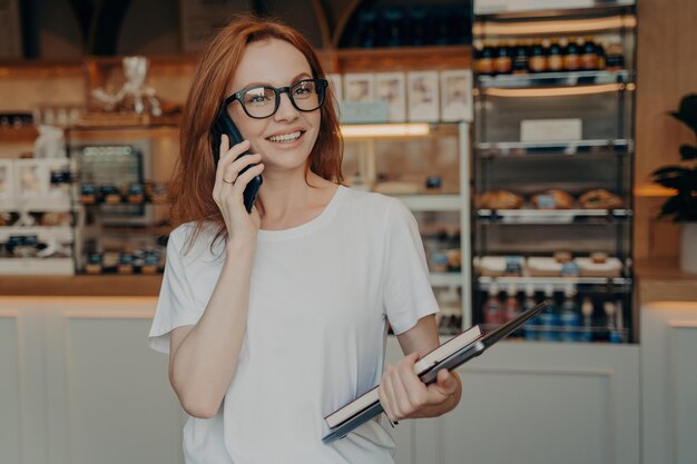 Cheerful young woman talking on the phone in a cafe holding a tablet with a casual look