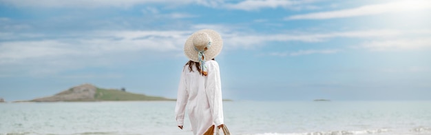 Cheerful young woman traveler with straw hat walking on beach during vacation back view