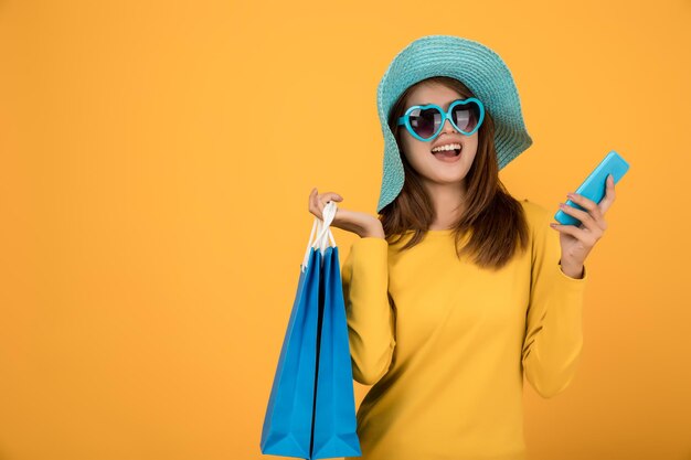 Cheerful young woman using mobile phone while holding shopping bags against yellow background