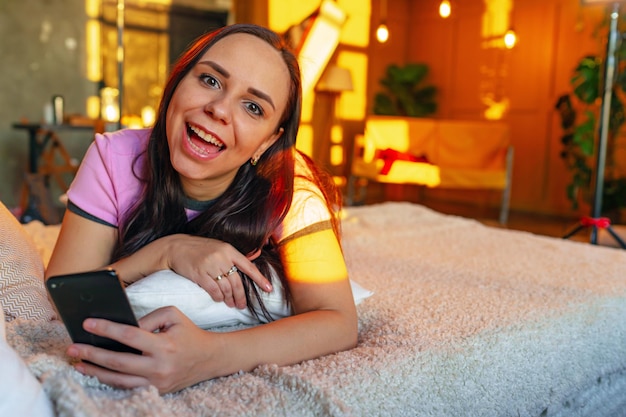 Cheerful young woman using smartphone on bed From above happy youthful female looking at camera while browsing on cellphone and lying on bed