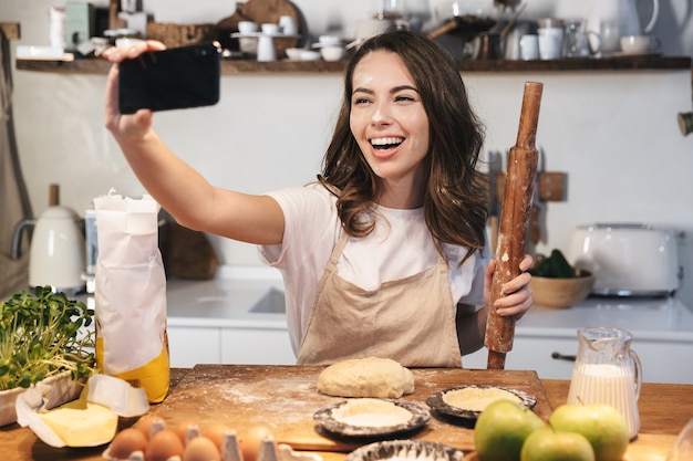 Cheerful young woman wearing apron preparing dough for an apple pie at the kitchen at home, taking a selfie, holding rolling pin