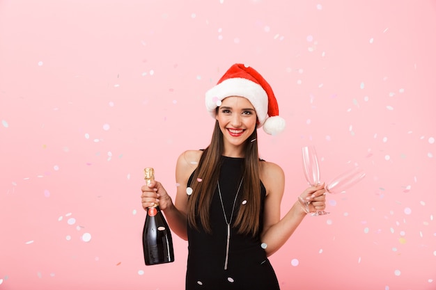 Cheerful young woman wearing Christmas hat celebrating standing isolated over pink background, holding bottle of champagne and glasses