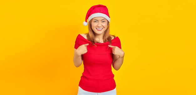 Cheerful young woman wearing Christmas hat pointing to herself on yellow background