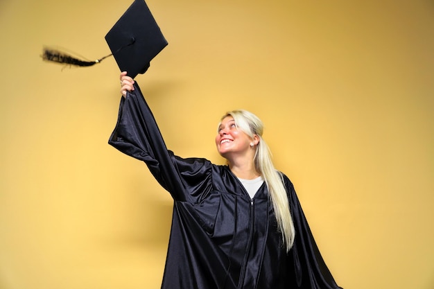 Foto giovane allegra che indossa l'abito di laurea mentre tiene il cemento contro uno sfondo giallo