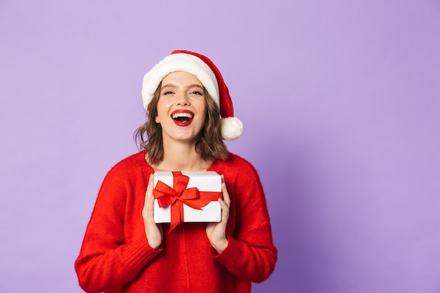 Cheerful young woman wearing red Christmas hat standing isolated over violet wall, holding gift box