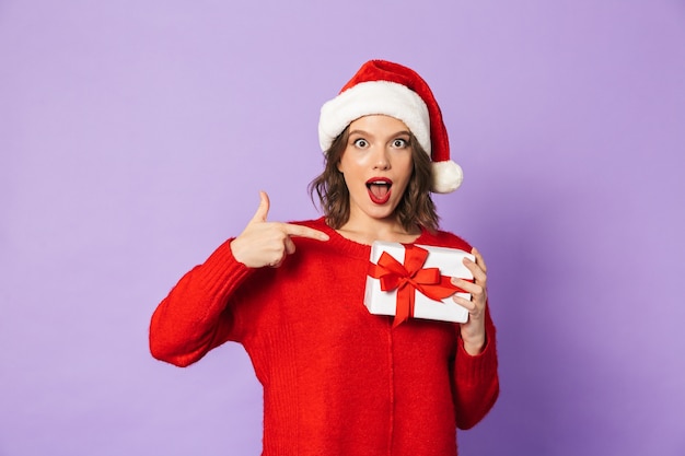 Giovane donna allegra che indossa il cappello rosso di natale in piedi isolato sul muro viola, tenendo il contenitore di regalo