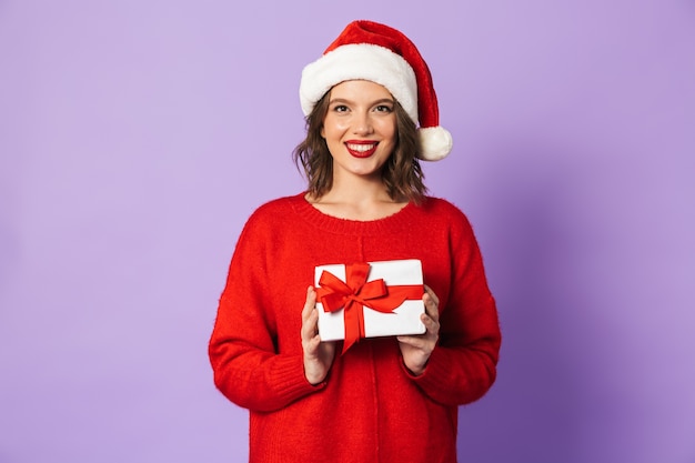 Cheerful young woman wearing red Christmas hat standing isolated over violet wall, holding gift box