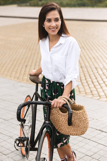 Cheerful young woman wearing summer clothes walking on a city street with bicycle