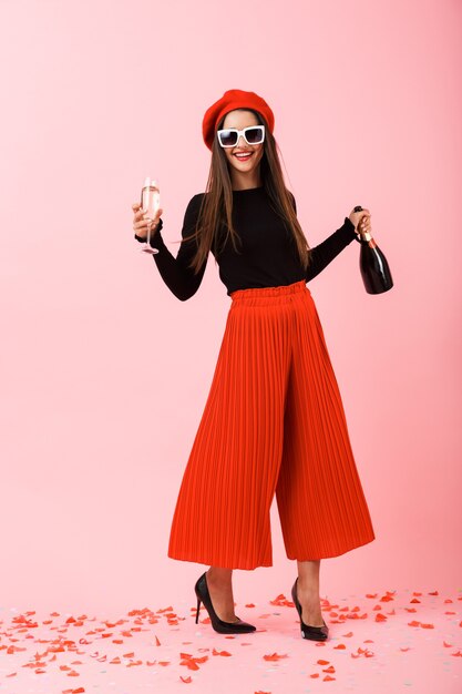 Cheerful young woman wearing sunglasses and red beret celebrating, holding bottle of champagne and a glass