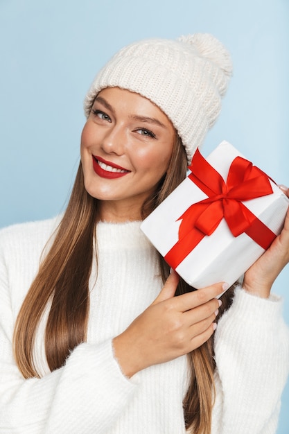 Cheerful young woman wearing sweater and hat, holding gift box