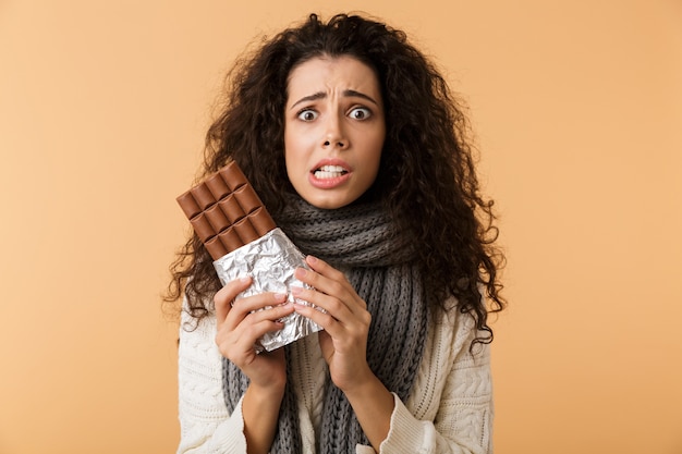 Cheerful young woman wearing sweater and scarf holding big chocolate bar isolated over beige wall