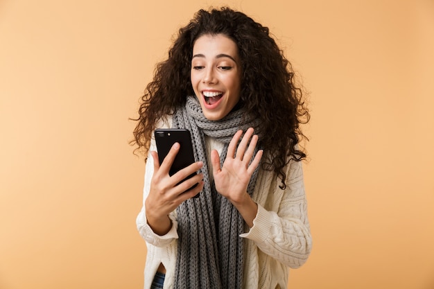 Cheerful young woman wearing winter scarf standing isolated over beige wall, holding mobile phone, waving