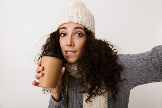 Cheerful young woman wearing winter scarf standing isolated over white wall, taking a selfie, holding a takeaway coffee cup