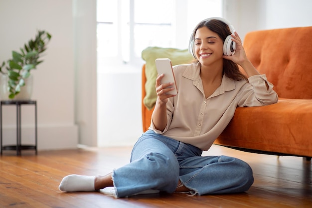 Cheerful young woman wearing wireless headphones sitting on the floor listening music