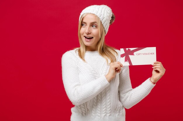 Cheerful young woman in white sweater, hat isolated on red wall background, studio portrait. Healthy fashion lifestyle, cold season concept. Mock up copy space. Hold gift certificate, looking aside.