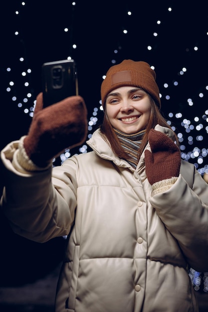 Cheerful young woman in winter clothes having video call on smartphone