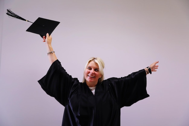 Cheerful young woman with arms raised standing in graduation gown against pink background