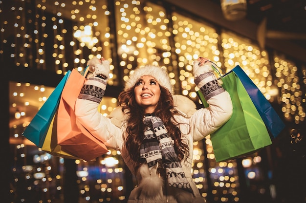 クリスマスの時期に街の通りで楽しんでいるカラフルな買い物袋を持つ陽気な若い女性。