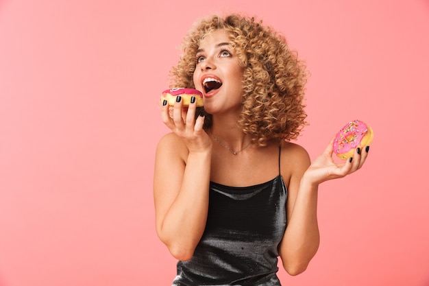 Photo cheerful young woman with curly hair, eating glazed donuts