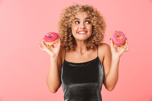 Cheerful young woman with curly hair, eating glazed donuts