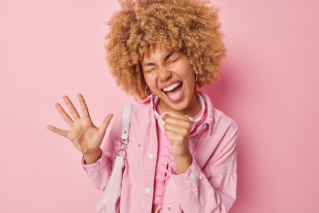 Cheerful young woman with curly hair keeps hand as if\
miscrophone sings song along wears stylish jacket carries bag has\
upbeat mood isolated over pink background people and emotions\
concept