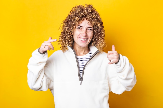 Cheerful young woman with curly hair shows gestures on a yellow background.