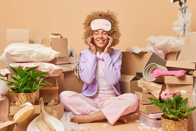 Cheerful young woman with curly hair sits crossed legs applies\
hydrogel patches under eyes going to celebrate relocation in new\
home dressed in domestic clothes surrounded by cardboard boxes