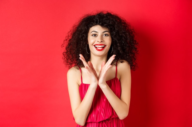 Cheerful young woman with curly hair, wearing evening dress, clap hands and smiling, praise good performance with applause, standing against red background.