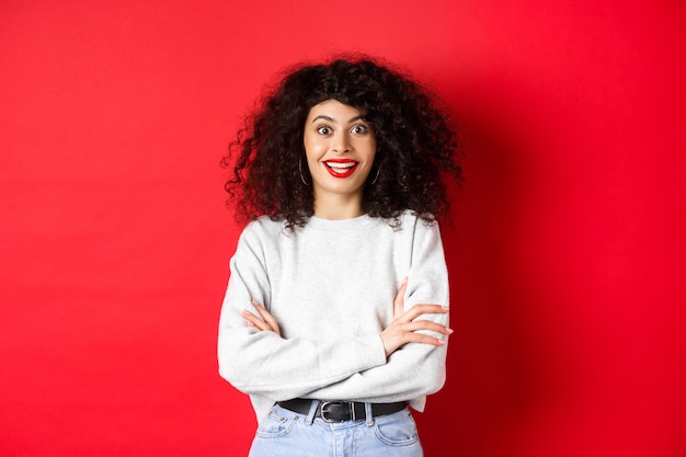 Cheerful young woman with curly hairstyle, raising eyebrows and looking surprised, hear interesting news, red background.