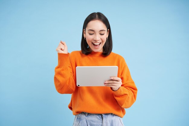 Cheerful young woman with digital tablet looks happy at gadget and celebrating laughing and person