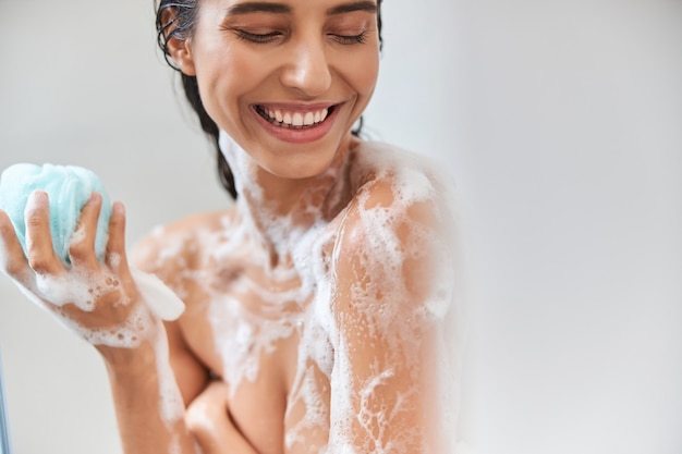 Photo cheerful young woman with foam on her body holding bath loofah