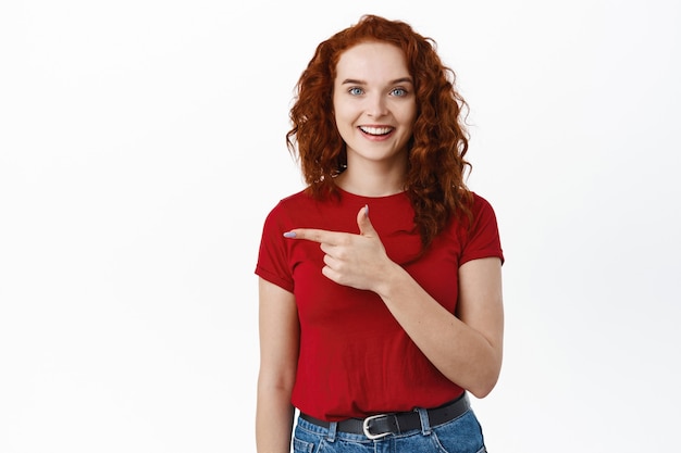 Cheerful young woman with ginger curly hair, pointing finger left and smiling happy , inviting check out promo deal on white wall