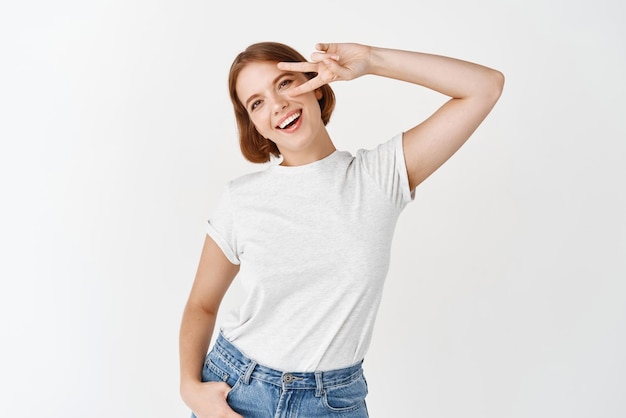 Cheerful young woman with short hair showing vsign over eye standing positive and happy white background Copy space