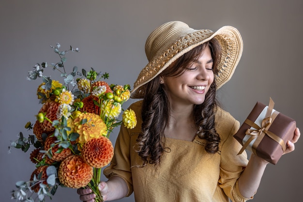 Cheerful young woman in a yellow dress and hat with a bouquet of bright chrysanthemums and a gift on a gray background.