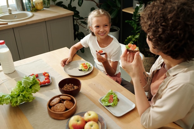 Cheerful youthful girl eating tasty sandwich and looking at her mother