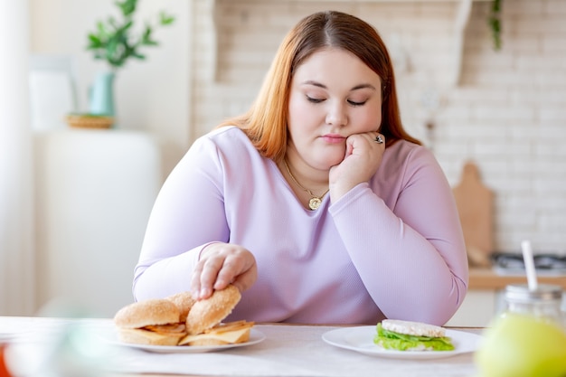 Безрадостная пухлая женщина смотрит на гамбургер, думая, есть его или нет